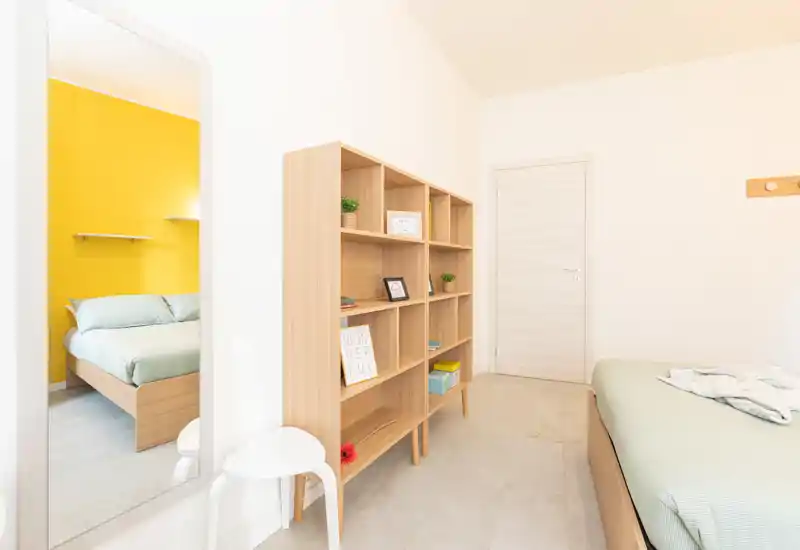 Chambre lumineuse et moderne avec mobilier en bois clair et un mur jaune en accent visible par la porte ; le lit est bien présenté avec une literie douce et une décoration soignée, parfaite pour une annonce locative.