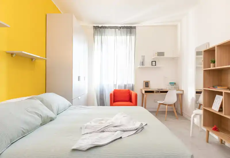 Chambre moderne et lumineuse avec un grand lit au premier plan, linge de lit menthe, mur jaune et un fauteuil orange près d'une fenêtre très lumineuse. Placard, bureau et étagères visibles — idéale pour un séjour confortable ou un logement étudiant.