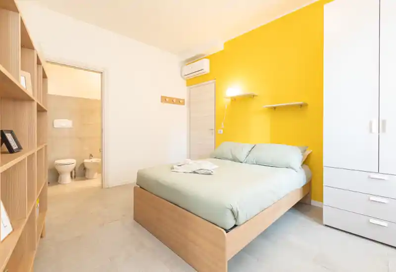 Chambre moderne et lumineuse avec un lit double en bois devant un mur jaune, une armoire blanche, des étagères ouvertes et une salle de bain attenante visible par la porte.