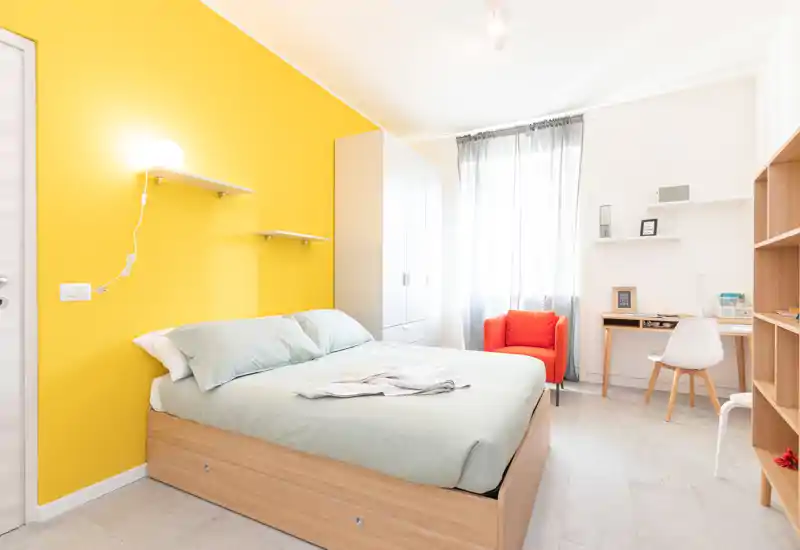 Chambre lumineuse et moderne avec lit double devant un mur d'accent jaune vif, armoire, bureau et un fauteuil orange confortable près de la fenêtre.