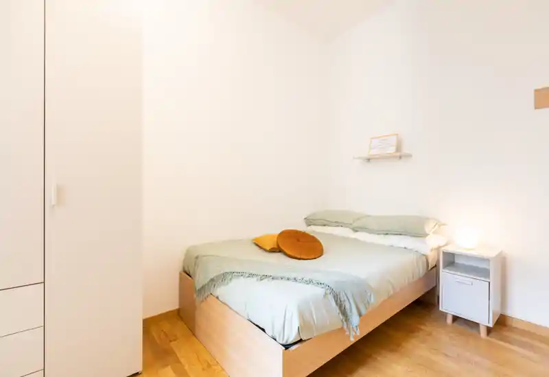 Camera da letto luminosa e minimal con letto matrimoniale in legno, biancheria verde chiaro e cuscini di accentuazione caldi; comodino con lampada e una mensola semplice che creano un ambiente accogliente.