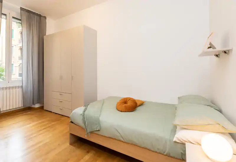 Camera da letto luminosa e minimal con letto singolo, lampada da comodino e armadio indipendente. La biancheria verde, il pavimento in legno e la finestra che illumina naturalmente rendono la stanza fresca e ordinata.