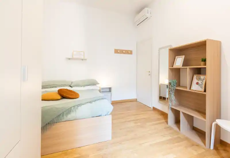 Camera da letto luminosa e minimal con struttura del letto in legno, biancheria verde tenue e cuscini decorativi; pavimento in legno caldo e scaffalatura ordinata per un ambiente accogliente e moderno.