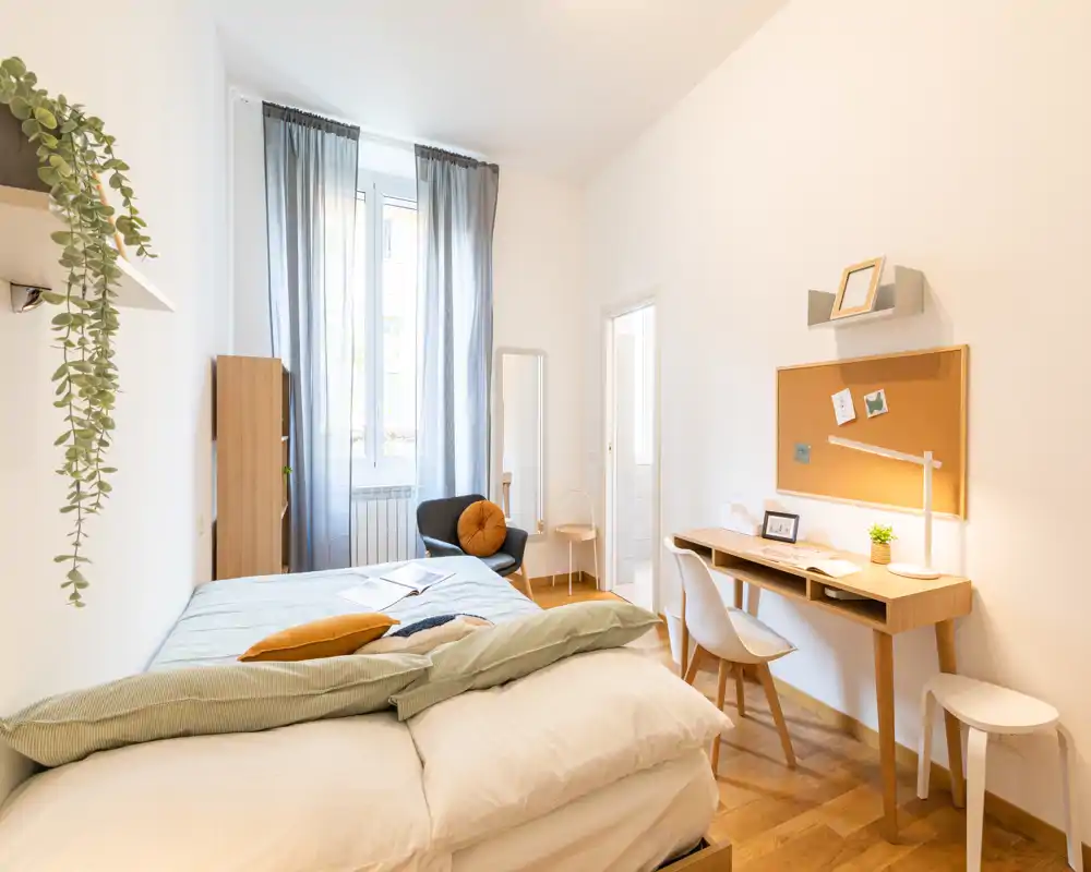 Chambre lumineuse et bien mise en scène avec un lit double confortable, un bureau équipé d’un panneau en liège et un fauteuil cosy près de la fenêtre — idéale pour une personne seule ou un couple.