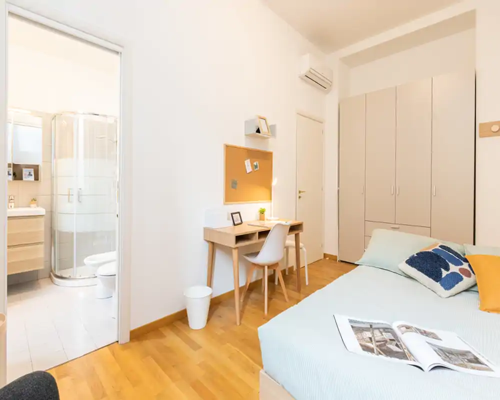 Chambre lumineuse et bien mise en scène avec un lit double, un bureau et une armoire intégrée ; salle de bains attenante avec douche visible.