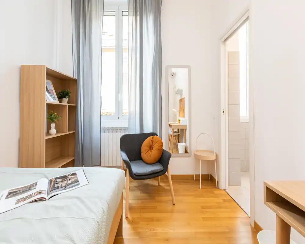 Chambre lumineuse et minimaliste avec lit au premier plan, fauteuil confortable avec coussin rond, étagères en bois et lumière naturelle provenant d'une grande fenêtre.