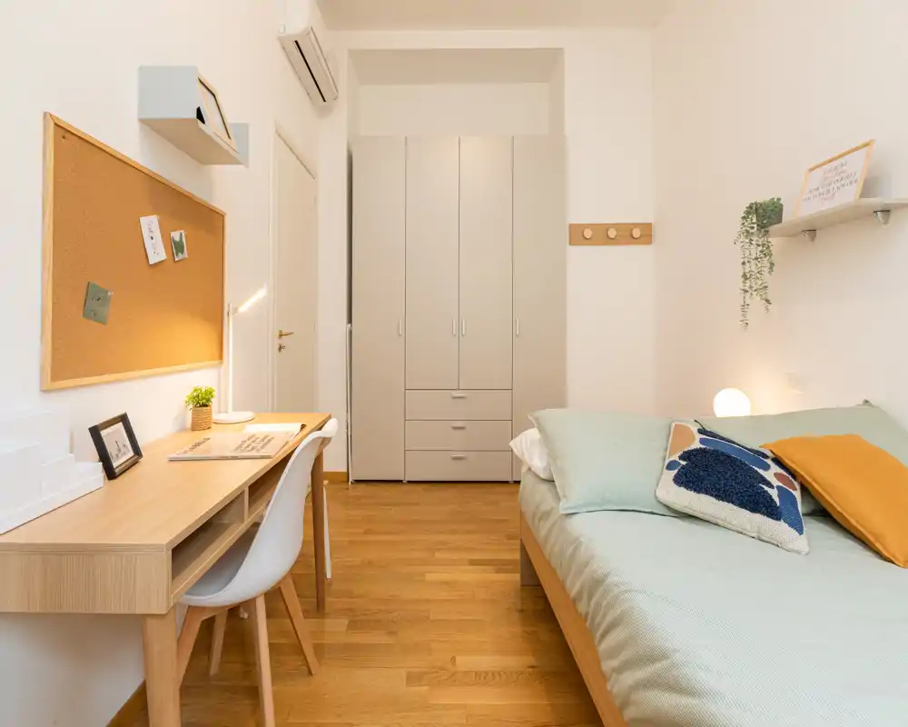Chambre simple lumineuse et bien mise en scène avec un lit en bois, coussins décoratifs et un bureau. Tons neutres et parquet chaleureux pour une ambiance moderne et apaisante, idéale pour la location longue durée.