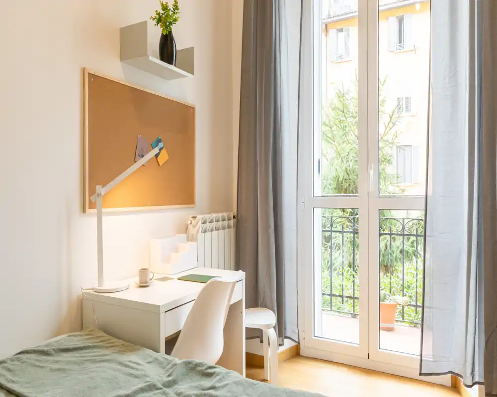 Camera luminosa e ordinata con letto singolo, scrivania e grande finestra con accesso al balcone — ideale per studenti o soggiorni brevi.