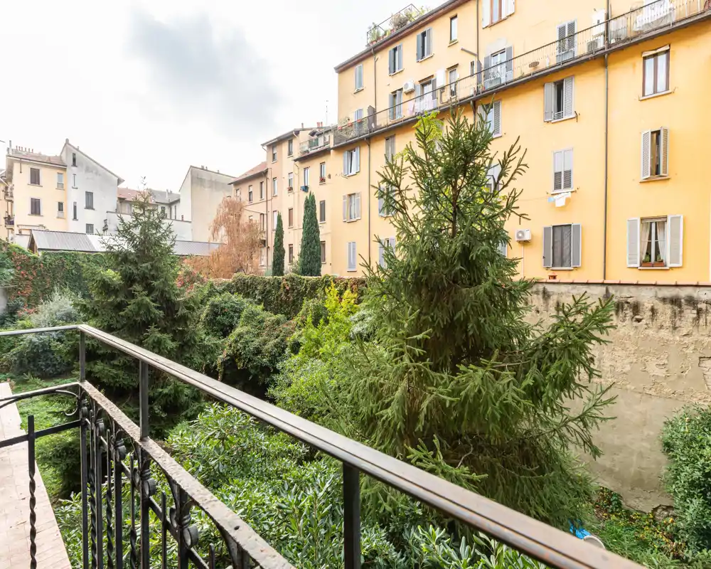 Vista dal balcone su un cortile verde con alberi maturi e le facciate degli appartamenti vicini — uno spazio esterno luminoso e ideale per rilassarsi o prendere un caffè.