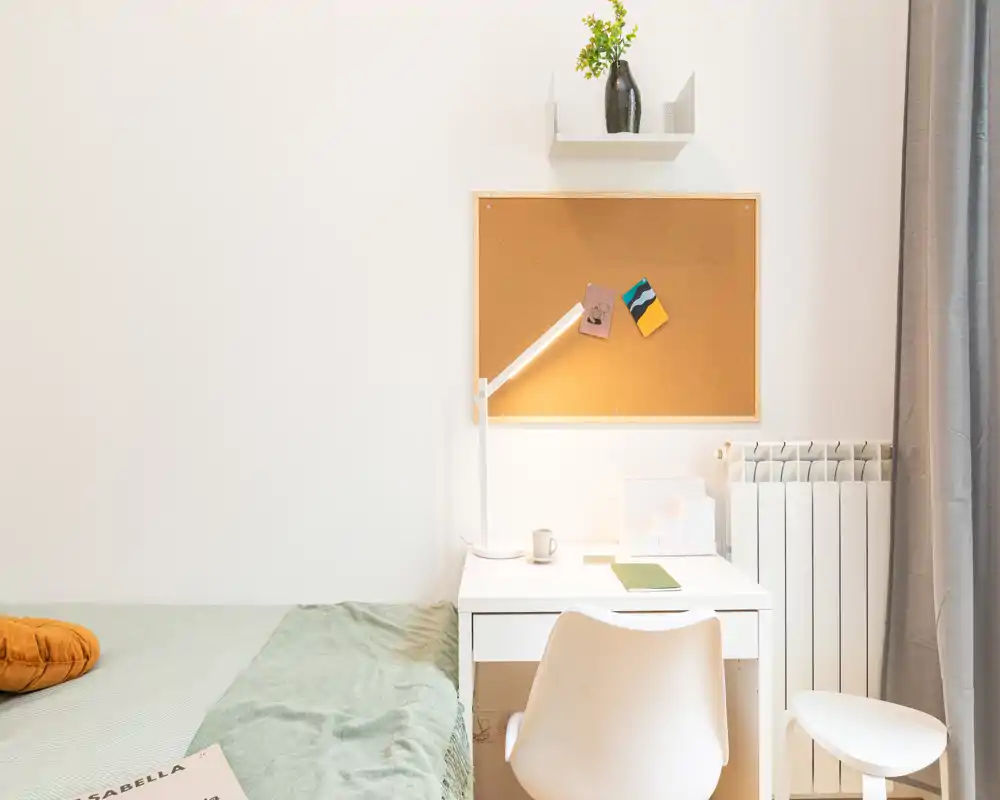 Angolo camera luminoso e minimale con letto ordinato, una scrivania bianca compatta e una lampada da tavolo calda — ideale per una persona singola.