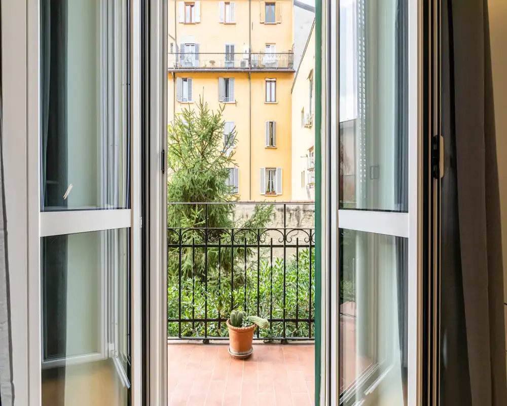 Doppie porte aperte che si affacciano su un piccolo balcone piastrellato con una pianta in vaso e una ringhiera in ferro, con vista su un cortile con verde e palazzi adiacenti.