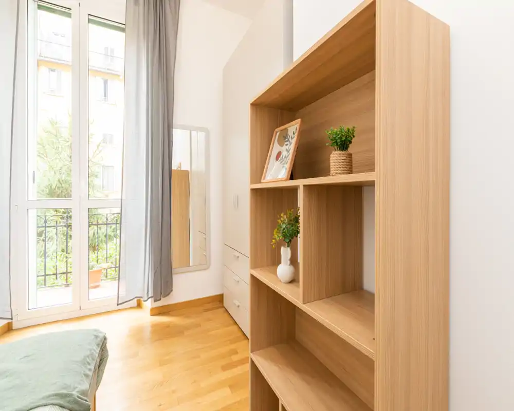 Angolo di camera da letto luminoso e minimale con libreria in legno, armadio, finestra a tutta altezza e una piccola porzione del letto visibile. Luce naturale e arredi semplici creano un ambiente arioso e tranquillo, ideale per annunci.