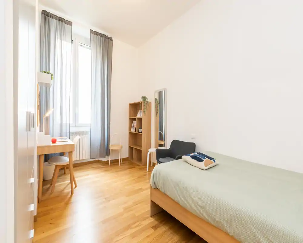 Camera singola luminosa e semplice, con letto in legno, scrivania vicino alla finestra, poltrona e libreria — parquet caldo e luce naturale soffusa.