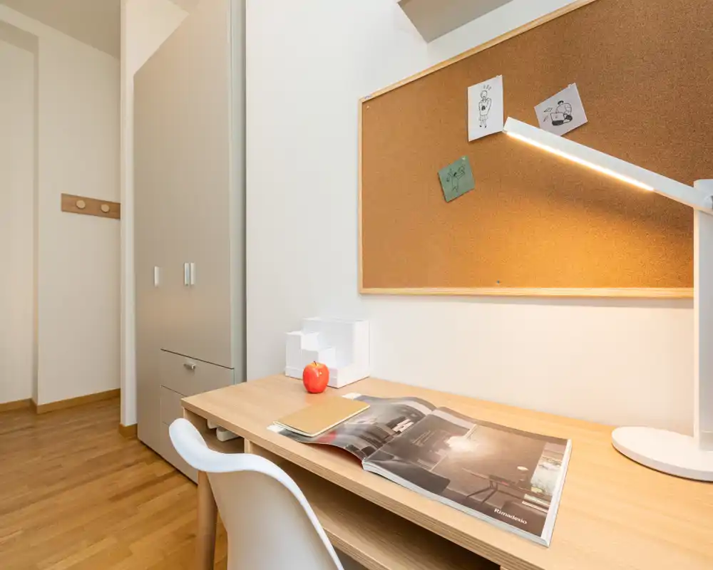 Angolo studio in camera da letto compatto con scrivania in legno, lampada da lavoro, bacheca in sughero e armadio — luminoso, ordinato e adatto allo studio.