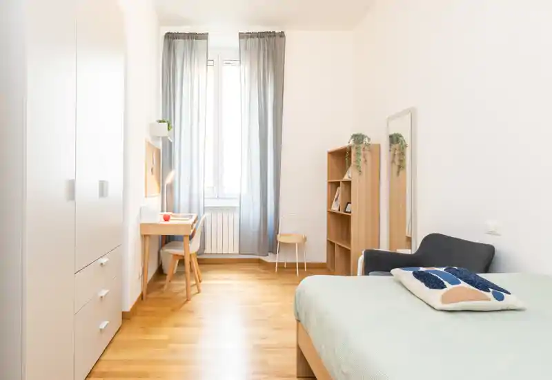 Bright, neatly staged bedroom with a single bed, comfortable chair, desk by the window and built-in wardrobe — ideal for a student or single occupant.