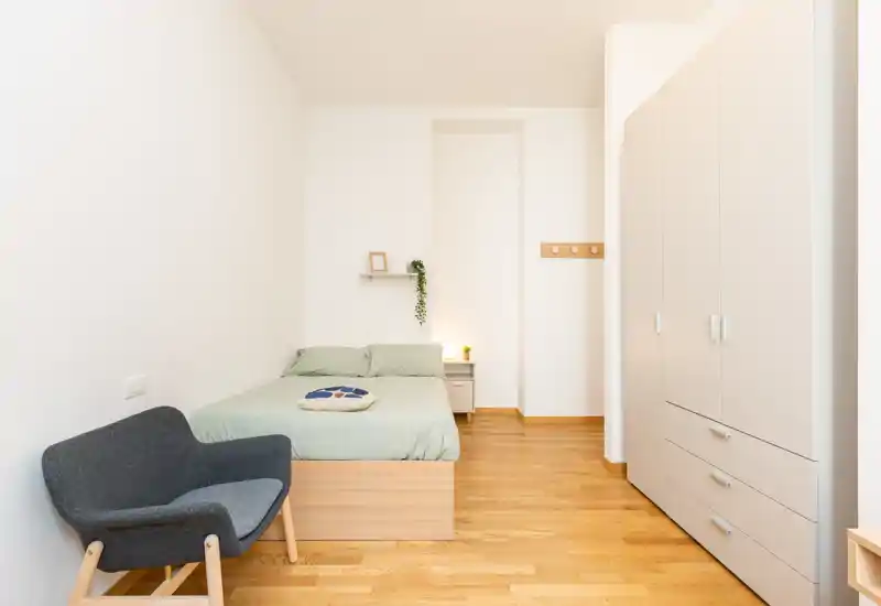 Bright, minimal bedroom with a double bed, bedside table, wardrobe and a comfortable accent chair — ideal for a tidy single or couple. Clean lines, warm wooden floor and soft natural light create a calm, welcoming atmosphere.