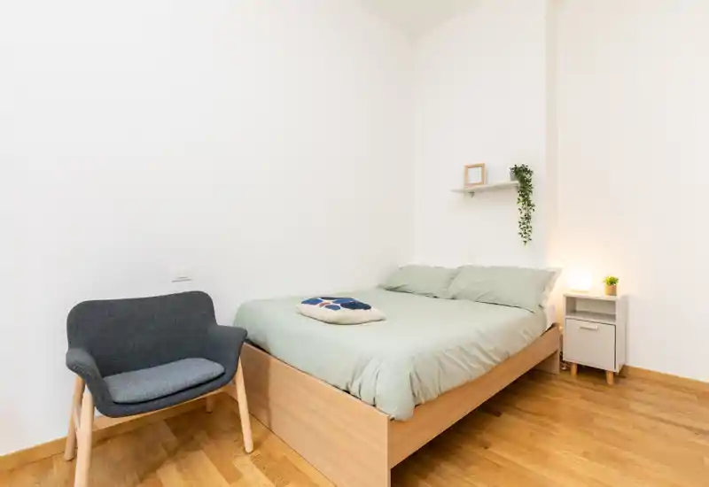 Bright, minimalist bedroom with a comfortable double bed, bedside cabinet with lamp and a small decorative plant, and a cozy accent chair — ideal for a calm, modern stay.