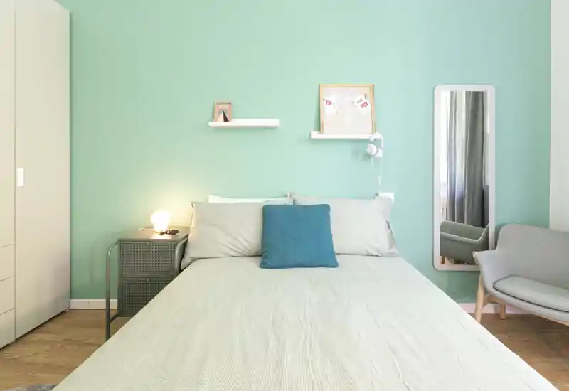 Bright, modern bedroom with a queen bed centered against a mint-green accent wall, bedside lamp, wall shelves and a full-length mirror — cozy and well-lit space ideal for rental listings.
