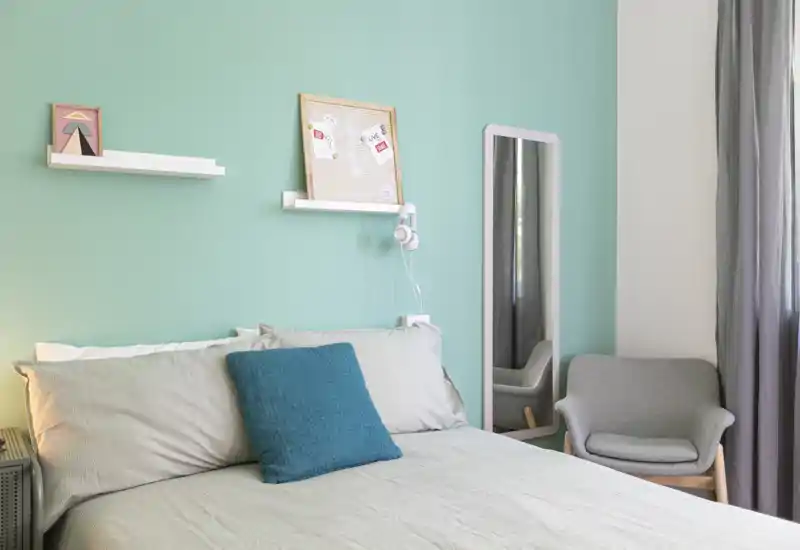 Well-staged bedroom with a neatly made double bed, teal accent pillow, wall-mounted decor and a cozy armchair by the window — bright, calm and inviting.