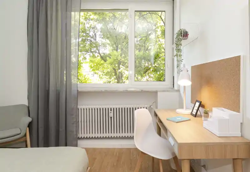 Angolo camera luminoso e ben organizzato con letto, una poltroncina confortevole e una scrivania in legno vicino alla finestra — ideale per uno studente o per il telelavoro.