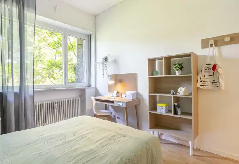 Bright, modern bedroom with a neatly made bed, a study desk by the window and open shelving. Large window provides abundant natural light and soft greenery view.