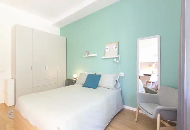 Bright, modern bedroom with a full-size bed, teal accent wall, wardrobe and cozy reading chair — ideal for calm stays.