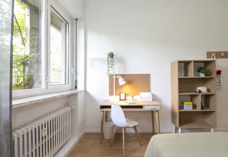 Angolo camera da letto luminoso e minimale con scrivania vicino a una grande finestra, libreria in legno e letto parzialmente visibile. Ideale per una persona singola che cerca luce naturale e uno spazio di lavoro ordinato.
