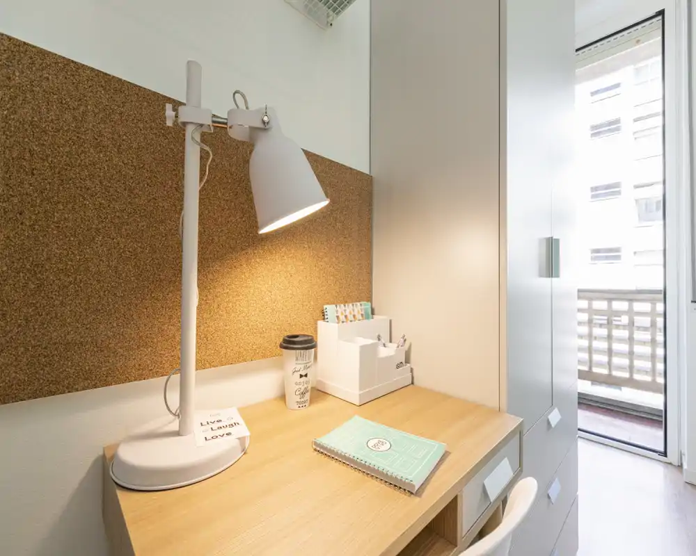Small, bright workspace next to a wardrobe and balcony access; wooden desk with a task lamp, cork board and stationery organised for study or remote work.