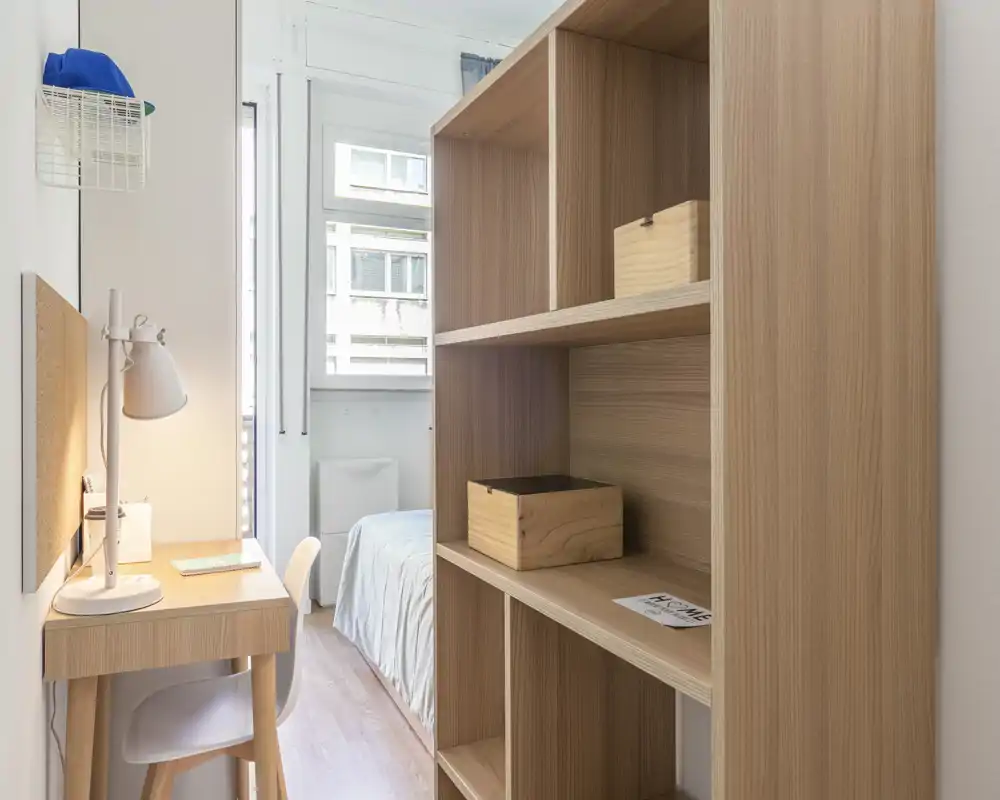 Compact, bright bedroom with a single bed visible through a doorway, natural light and wooden shelving creating a warm, tidy feel.