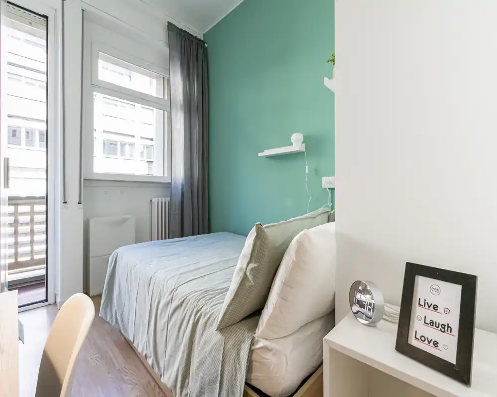 Bright, compact bedroom with a double bed against a teal accent wall, natural light from a window and balcony door, and simple, modern furnishings.
