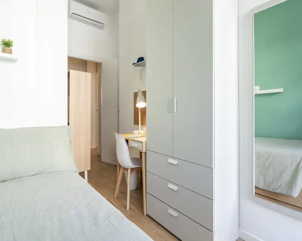 Bright, minimalist bedroom with a comfortable bed, a tall wardrobe, and a small desk with lamp — ideal for a single occupant or student.