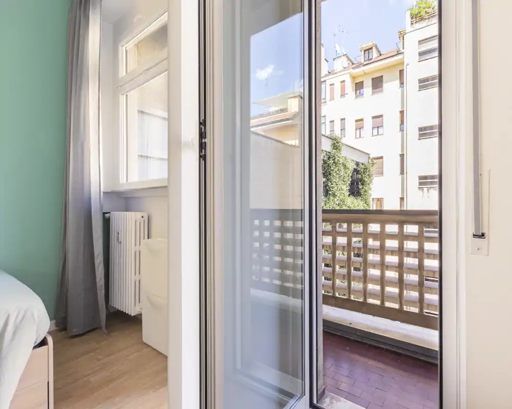 Bright small bedroom corner with a sliding glass door opening onto a balcony; light wooden floor, radiator and neutral textiles create a fresh, airy feel.