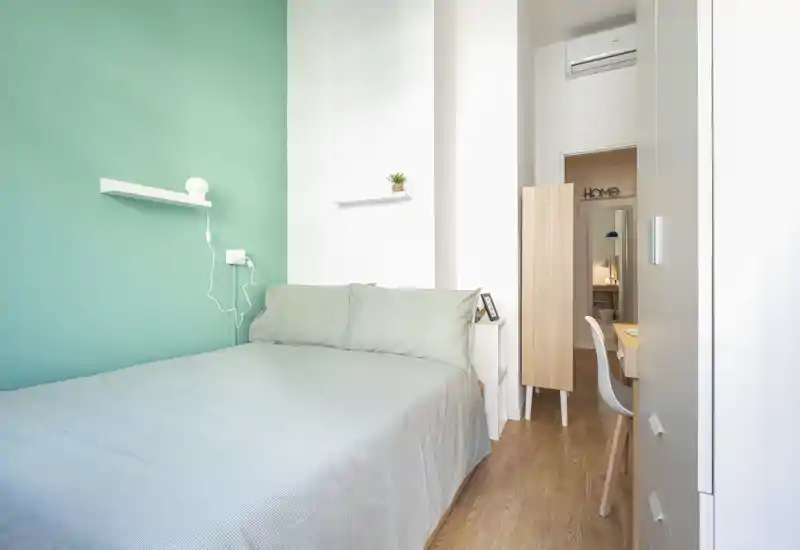 Bright, compact bedroom with a double bed against a teal accent wall, crisp linens and light wood flooring — ideal for a cozy, modern rental.