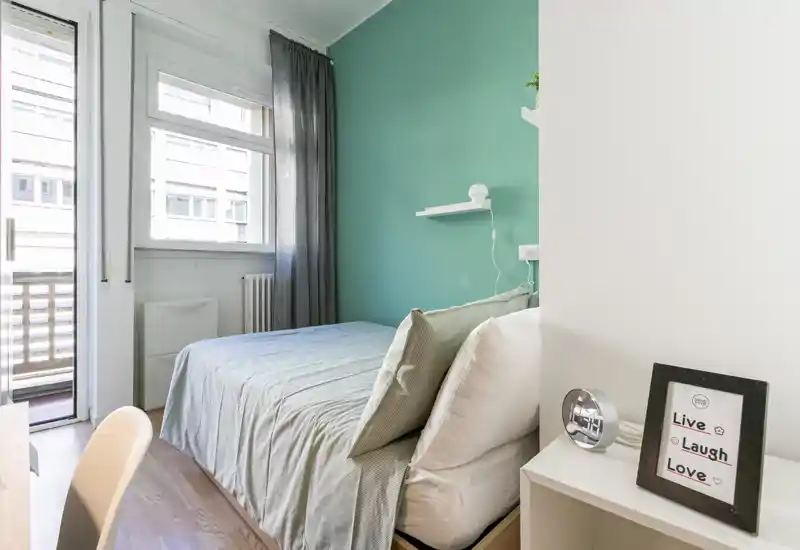 Bright, compact bedroom with a double bed against a teal accent wall, natural light from a window and balcony door, and simple, modern furnishings.