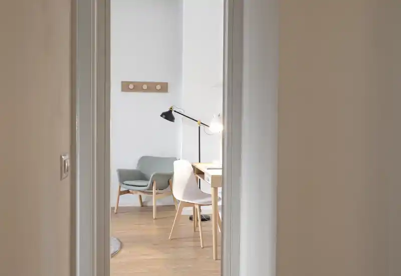 Espace salon chaleureux et bien éclairé vu depuis l’embrasure d’une porte, avec fauteuil, bureau et lampe — idéal pour un coin bureau ou un petit logement.