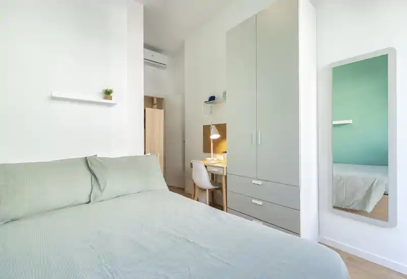 Bright, minimal bedroom with a large bed, built-in wardrobe and a small desk area; fresh, calm colors and neat staging ideal for rental listings.
