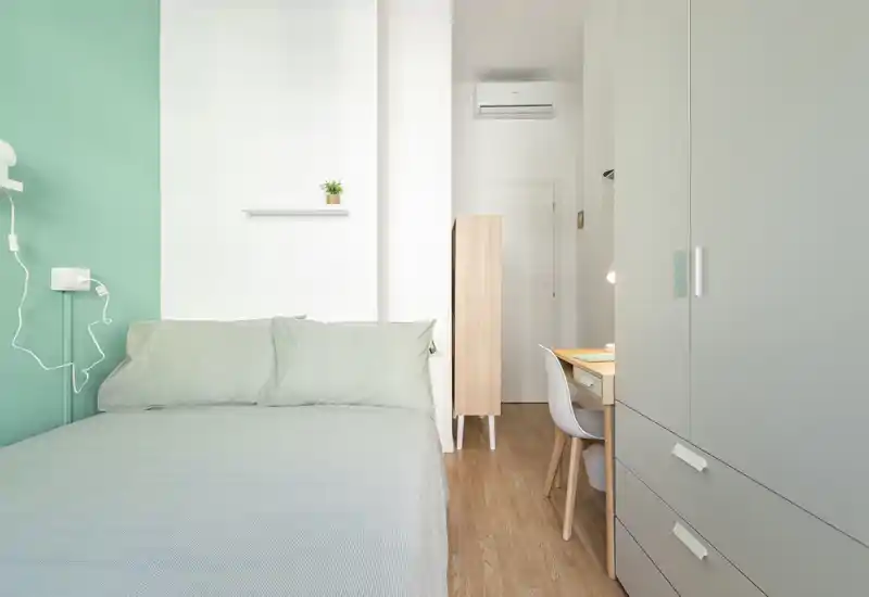 Bright, compact bedroom with a neatly made double bed, soft pastel bedding and a small desk beside a wardrobe — ideal for a single occupant or couple.