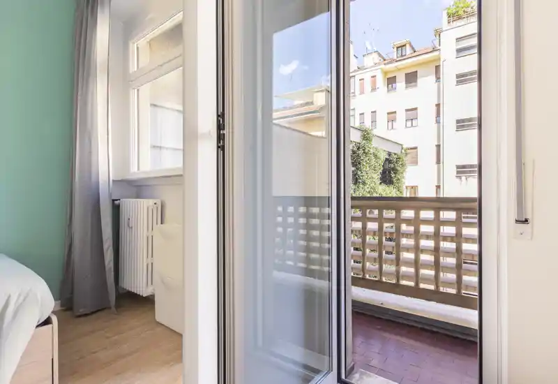 Coin chambre lumineux avec porte-fenêtre coulissante donnant sur un balcon ; parquet clair, radiateur et textiles neutres pour une ambiance aérée.