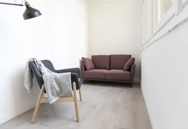 Piccola zona giorno luminosa con divano due posti compatto e una poltrona con coperta; ambiente minimal e ben illuminato, perfetto come angolo lettura accogliente.