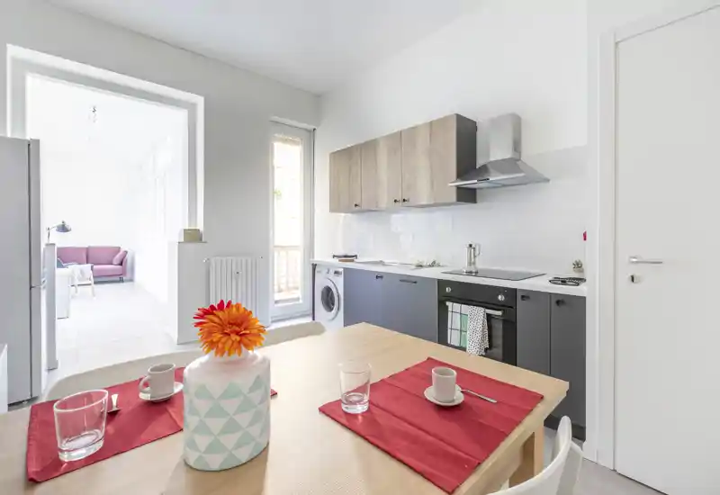 Cucina moderna e luminosa con elettrodomestici integrati e piccolo tavolo da pranzo per due; luce naturale dal balcone e stile minimale e curato.