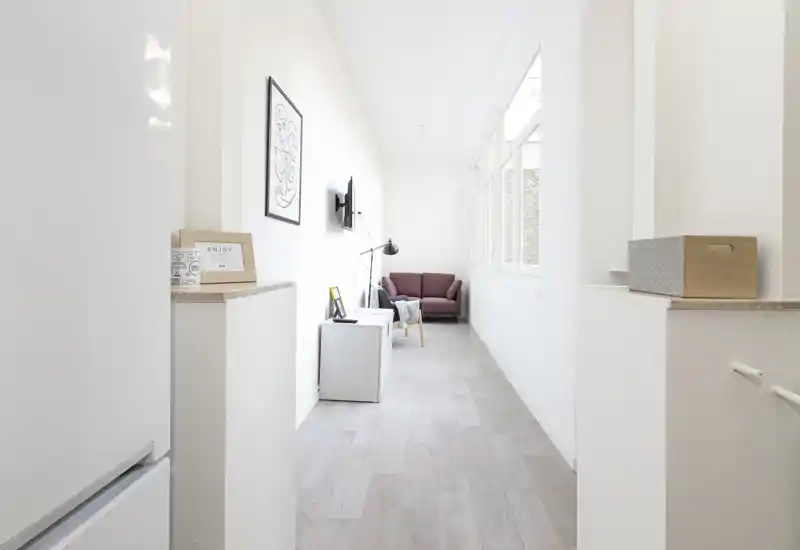 Zona living luminosa e lunga con poltrona color bordeaux, lampada da terra e TV a parete — stile minimale e ampie finestre che garantiscono molta luce naturale.
