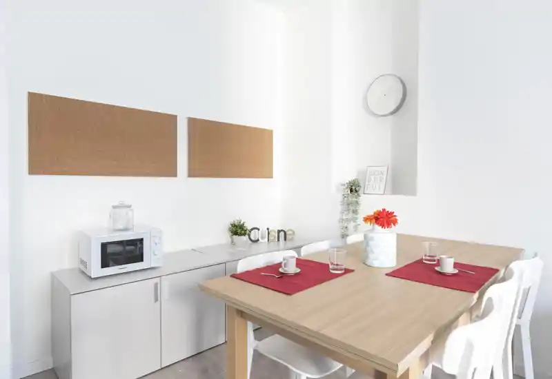 Zona pranzo moderna e luminosa con tavolo in legno apparecchiato per due, sedie bianche e dettagli decorativi come vaso con fiori e pannelli in sughero a parete.