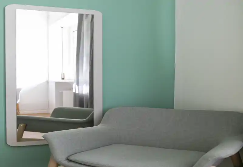 Angolo living compatto e di design con divano imbottito in stile mid-century su parete verde menta e specchio a tutta altezza che riflette luce naturale.