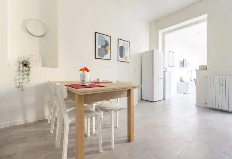 Zona pranzo luminosa e minimal con tavolo in legno, sedie bianche e quadri decorativi; la luce naturale dalla stanza adiacente rende l'ambiente arioso e accogliente.