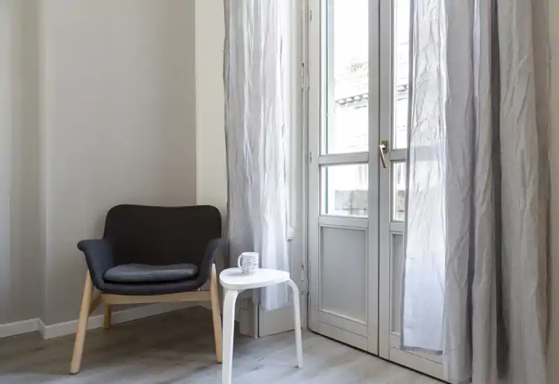 Coin salon lumineux et minimaliste avec un fauteuil confortable, une petite table d'appoint blanche et des rideaux légers devant une porte-fenêtre — un endroit calme pour lire ou se détendre.