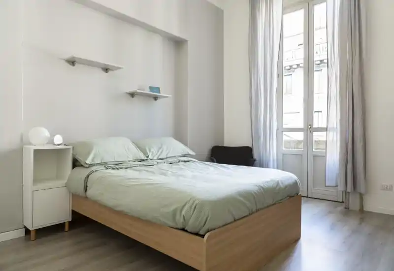 Chambre lumineuse et minimaliste avec lit en bois, linge de lit vert pâle, table de chevet et grande fenêtre laissant entrer beaucoup de lumière naturelle.