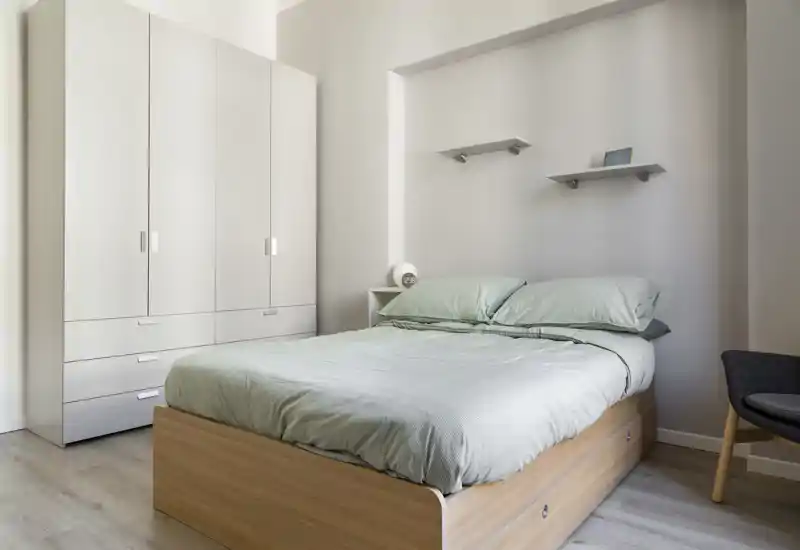 Chambre lumineuse et minimaliste avec un lit double sur cadre en bois clair et une grande armoire blanche ; linge de lit vert pâle et étagères simples pour une ambiance moderne et apaisante.