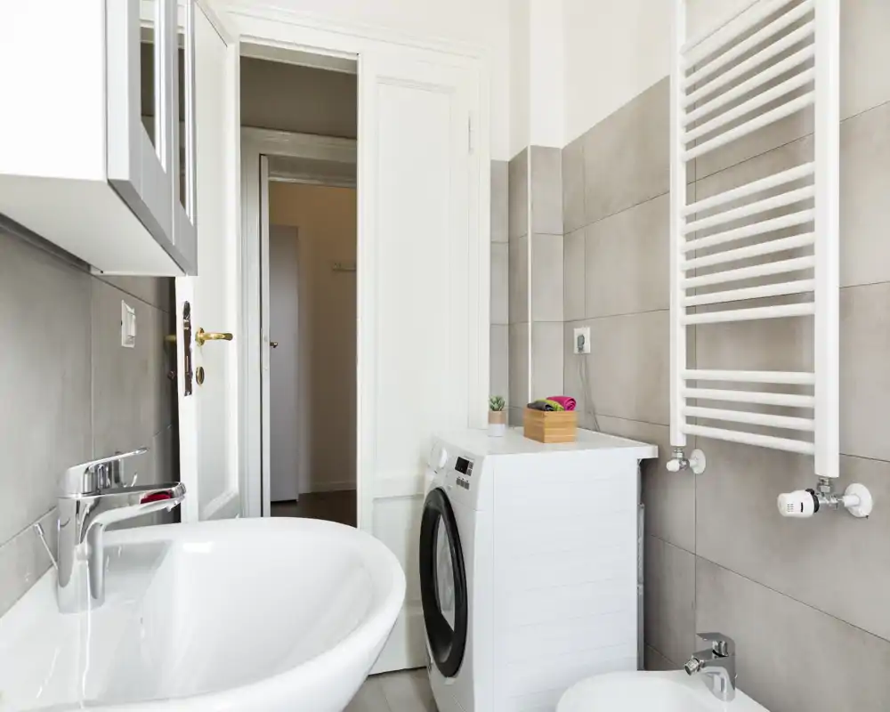 Salle de bains moderne et lumineuse avec lavabo, bidet, radiateur porte-serviettes mural et machine à laver à côté du lavabo ; carrelage gris clair et style épuré.