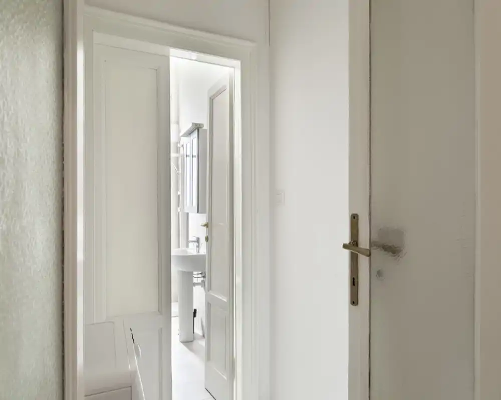 Salle de bains lumineuse et minimaliste vue à travers une porte ouverte, avec lavabo sur colonne et armoire miroir dans un décor blanc épuré.