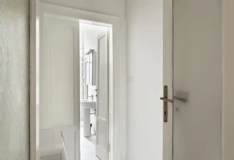 Bright, minimalist bathroom seen through an open doorway featuring a pedestal sink and mirrored cabinet in a clean white palette.